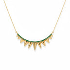 A gold necklace with a curved row of sharp spike charms, accented by a line of small green gemstones along the top curve, shown on a plain white background.