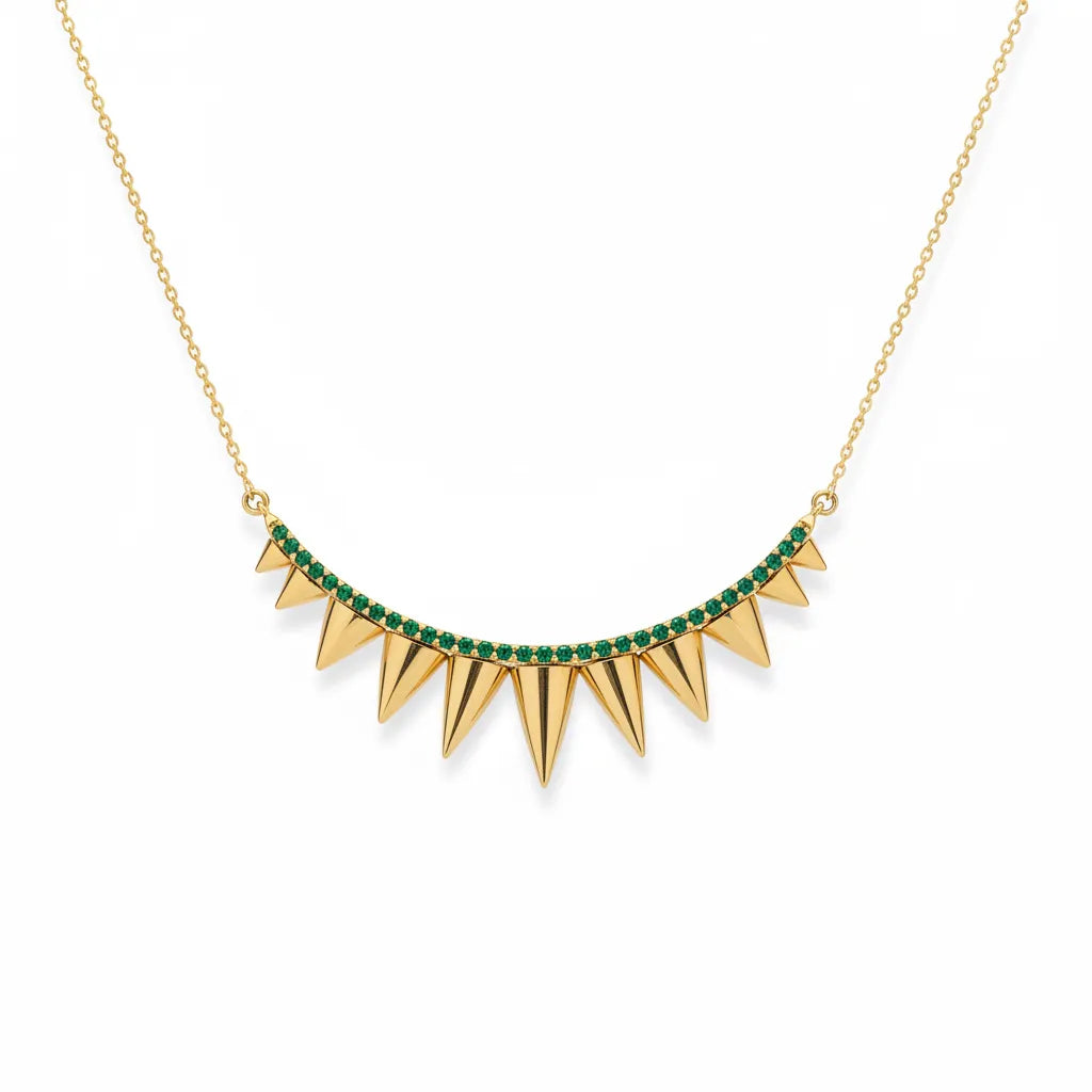 A gold necklace with a curved row of sharp spike charms, accented by a line of small green gemstones along the top curve, shown on a plain white background.