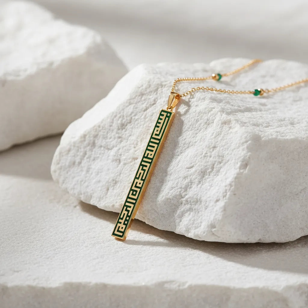 A gold bar pendant necklace with a green Greek key pattern, resting on a textured white stone against a soft white background.