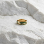 A gold band ring with a green Greek key pattern, resting on a textured white stone surface.